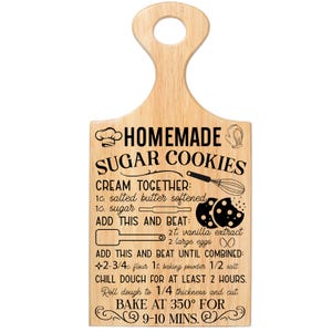 May include: Wooden cutting board with a handle, featuring a recipe for homemade sugar cookies. The text includes ingredients and baking instructions, such as "Bake at 350&deg; for 9-10 mins." The board has a light wood grain.
