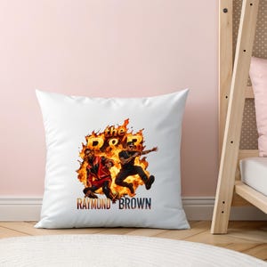 Pu&ograve; includere: Un cuscino quadrato bianco con una stampa grafica di due figure davanti a fiamme arancioni e gialle. Il testo "the R&B" appare sopra le figure e "RAYMOND BROWN" &egrave; stampato in grassetto nella parte inferiore del cuscino.