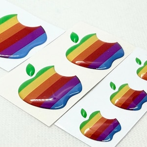 Peut inclure: Plusieurs autocollants avec un motif de pomme rayée arc-en-ciel et une feuille verte. Les autocollants sont sur du papier blanc et de différentes tailles. Le motif de la pomme a une bouchée.