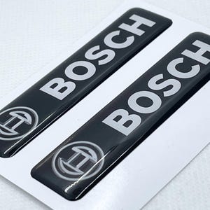 Puede incluir: Dos pegatinas negras con el emblema de Bosch en letras blancas y un logotipo blanco. Las pegatinas rectangulares tienen un acabado brillante. El logotipo de Bosch es una letra "H" estilizada dentro de un círculo.