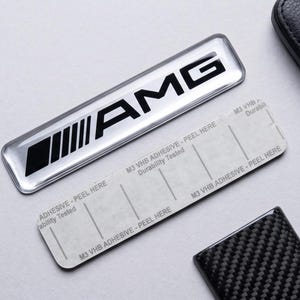 Könnte beinhalten: Ein rechteckiges, silber-schwarzes AMG-Emblem mit metallischer Oberfläche, begleitet von einem rechteckigen Klebestreifen mit der Aufschrift M3 VHB ADHESIVE - PEEL HERE, einem schwarzen, strukturierten Schlüsselanhänger und einer Pinzette.