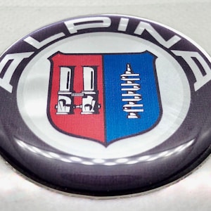 Może przedstawiać: Okrągła odznaka z napisem "ALPINA" w białych literach na zewnętrznej krawędzi. W środku tarcza podzielona na czerwone i niebieskie sekcje, z białymi i srebrnymi szczegółowymi grafikami.