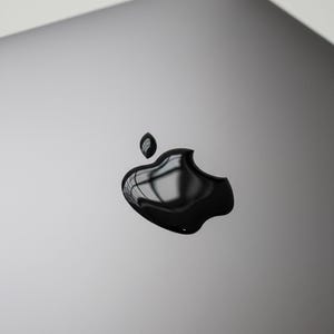 Könnte beinhalten: Nahaufnahme eines silbernen Laptops mit glänzendem schwarzem Apple-Logo. Das Logo reflektiert das Licht und erzeugt einen glänzenden Effekt. Die Oberfläche des Laptops ist glatt und sauber, mit minimalistischem Design.