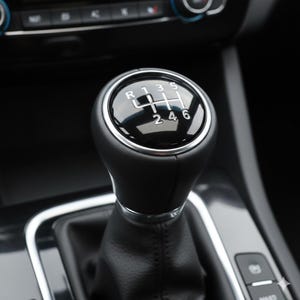 Könnte beinhalten: Nahaufnahme eines schwarzen Lederschaltknaufs mit einem Chromzierring. Das Schaltschema ist mit weißen Zahlen und Buchstaben sichtbar. Der Innenraum des Autos ist im Hintergrund zu sehen.