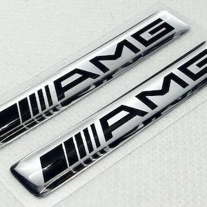 Pode incluir: Dois emblemas AMG prateados com letras pretas e um design de listras pretas e brancas. Os emblemas são retangulares e têm um acabamento brilhante. O logotipo AMG é exibido de forma proeminente em cada emblema.