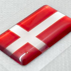 Puede incluir: Una pegatina con la bandera danesa, roja y blanca. La bandera es una cruz blanca sobre fondo rojo.