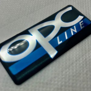 Könnte beinhalten: Ein silbernes und blaues 3D-Emblem mit dem Text "OPC LINE" auf schwarzem Hintergrund.