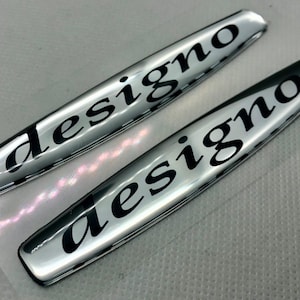 Op de afbeelding: Twee zilverkleurige auto-emblemen met het woord "designo" in zwarte letters.