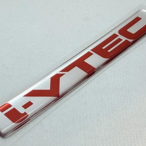 Könnte beinhalten: Ein rechteckiges silbernes i-VTEC-Emblem mit roter Schrift. Das Emblem befindet sich in einer transparenten Kunststoffhülle. Das i-VTEC-Logo ist ein Autoteil.