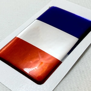 Op de afbeelding: Een rechthoekig, glanzend, driedimensionaal embleem met een horizontaal driekleurig ontwerp, bestaande uit een donkerblauw bovengedeelte, een wit middengedeelte en een felrood ondergedeelte op een witte rechthoekige rand.