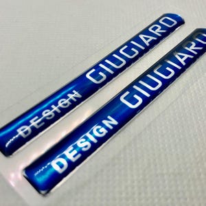 May include: Two blue and silver stickers with the text "DESIGN GIUGIARO" on each. The stickers are on a white background.
