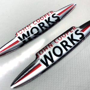 Puede incluir: Dos emblemas John Cooper Works plateados y rojos. Los emblemas tienen forma de una hoja larga y delgada con una franja roja en el centro. El texto "John Cooper Works" está impreso en negro en los emblemas.