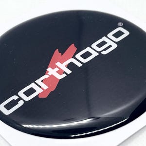 Pode incluir: Uma caneca de cerâmica branca com uma alça preta e o texto "But First, Coffee" em negrito preto. A caneca está sobre um fundo desfocado, sugerindo um ambiente acolhedor.