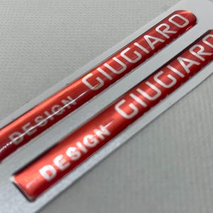 Op de afbeelding: Twee rode Giugiaro Design stickers met witte tekst. De stickers zijn rechthoekig met afgeronde uiteinden en staan op een witte achtergrond. Het woord "DESIGN" staat aan de linkerkant van de sticker en "GIUGIARO" staat aan de rechterkant.