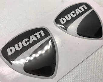 May include: Two black and white stickers with the word "DUCATI" in white letters on a black background. The stickers are shaped like a rounded triangle.