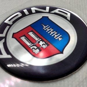 Op de afbeelding: Een ronde Alpina embleemsticker met een donkerblauwe buitenring en zilverkleurige tekst. Het midden toont een schild verdeeld in twee delen: een rode zijde met een zilverkleurige carburateurafbeelding en een blauwe zijde met een krukasafbeelding.