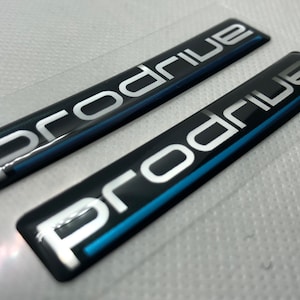 Könnte beinhalten: Zwei schwarze und silberne "Prodrive" Embleme mit einem blauen Streifen. Das obere Emblem ist leicht erhöht und das untere Emblem ist flach.