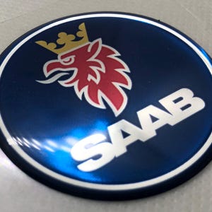 May include: A blue and white round emblem with a silver Saab logo and a red and white griffin with a gold crown.
