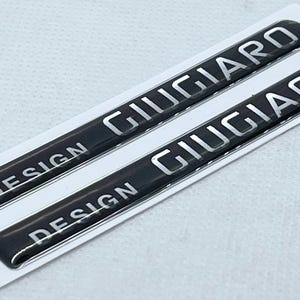 May include: Two black Giugiaro Design automotive emblems with silver lettering. The emblems are rectangular and slightly curved, with the word "DESIGN" at the top and "GIUGIARO" below. They are on a white backing.