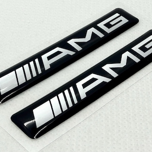Puede incluir: Dos emblemas AMG rectangulares negros con letras plateadas y un diseño de rayas plateadas. Los emblemas son brillantes y tienen una ligera curva. El logotipo AMG se muestra de forma destacada.