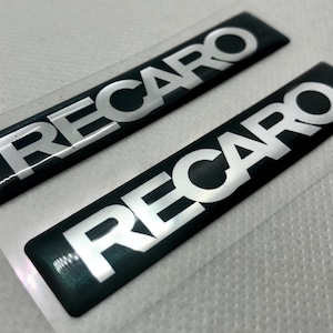 Op de afbeelding: Twee zwarte en zilveren Recaro auto-emblemen. De emblemen zijn rechthoekig en hebben een glanzende afwerking.