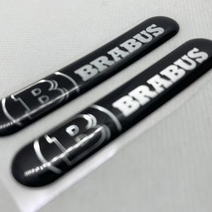 Puede incluir: Dos emblemas negros de forma ovalada con la palabra "BRABUS" en letras plateadas. Cada emblema también presenta un logotipo "B" estilizado. Los emblemas son brillantes y parecen estar hechos de un material duradero.