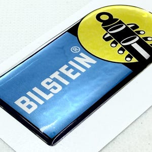 Peut inclure: Un autocollant rectangulaire brillant avec un fond bleu et le mot blanc "BILSTEIN". Le coin supérieur droit présente un cercle jaune avec un graphique noir et blanc. L'autocollant a une bordure noire et est sur une surface blanche.