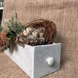May include: A rustic, white-painted wooden drawer with a round knob, holding a decorative bird's nest. The nest contains speckled eggs and is surrounded by dried flowers and greenery, creating a natural, farmhouse aesthetic.