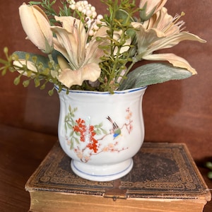 May include: A white ceramic vase with a floral design, filled with beige lilies and greenery. The vase features a painted bird and floral branch motif. The vase is placed on top of a vintage book with gold-edged pages.