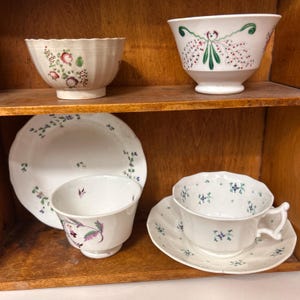 May include: A collection of antique white porcelain tableware with floral designs. The set includes bowls, plates, teacups, and saucers. The floral patterns feature red, green, and purple accents. The items are displayed on wooden shelves.
