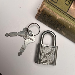 May include: A vintage silver-colored padlock with the text "Slaymaker Rustless" and two matching keys on a ring. A worn, green book is in the background.