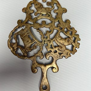 May include: Ornate brass trivet with a handle and openwork design. The trivet features a lion, a horse, and a bell. The brass has a golden hue and is likely vintage.