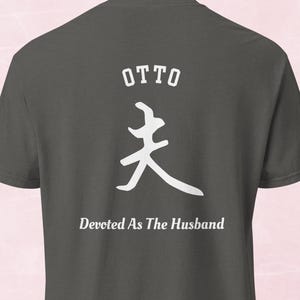 May include: Dark gray t-shirt with white text and a symbol. The word "OTTO" is above a white symbol, with the phrase "Devoted As The Husband" below. The shirt is made of a soft material.
