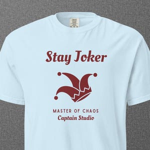 May include: Light blue t-shirt with the text "Stay Joker" above a maroon jester hat graphic. Below the graphic, the shirt reads "MASTER OF CHAOS" and "Captain Studio". The shirt is a short-sleeved crew neck.