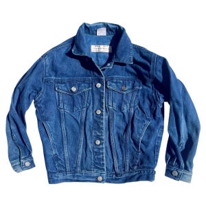 May include: A medium-wash blue denim jacket featuring a classic collar, long sleeves with buttoned cuffs, and a front button closure. The jacket includes two chest pockets with buttoned flaps and a white rectangular brand label at the inner neckline.