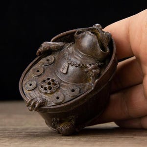 Puede incluir: Una escultura de rana de bronce con una base en forma de moneda y un dise&ntilde;o de flor de loto. La rana lleva un collar y est&aacute; sentada en un cuenco. La escultura se sostiene en una mano.