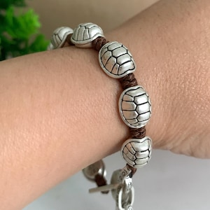 Può includere: Un braccialetto caratterizzato da una serie di ciondoli color argento a forma di guscio di tartaruga infilati su un cordino marrone scuro. I ciondoli sono distanziati da nodi sul cordino e il bracciale include una chiusura a T argentata.
