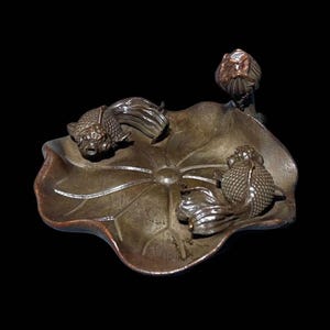 May include: A detailed metal sculpture of two goldfish swimming on a lily pad with a flower bud. The sculpture is in shades of silver and bronze, set against a black background. The fish have textured scales and flowing fins.