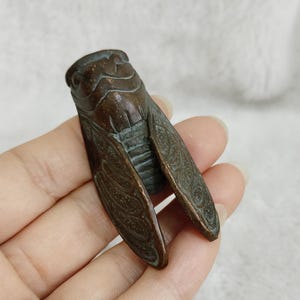 Könnte beinhalten: Bronze-farbene Zikadenfigur mit detaillierten Flügelmustern. Die Insektenskulptur hat eine strukturierte Oberfläche. Der Körper der Zikade ist dunkler Bronze, die Flügel heller. Die Figur wird in einer Hand gehalten.