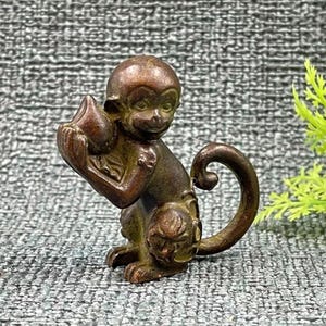 May include: A bronze monkey figurine, seated with a curled tail and holding a fruit-like object. The monkey has detailed facial features and is posed on a textured surface. The figurine is a decorative item.