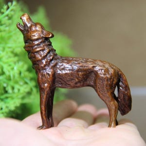 May include: A detailed, brown figurine of a wolf howling, with its head tilted back and mouth open. The wolf is standing, with its tail slightly raised. The figurine has a textured surface and is set against a blurred green and brown background.