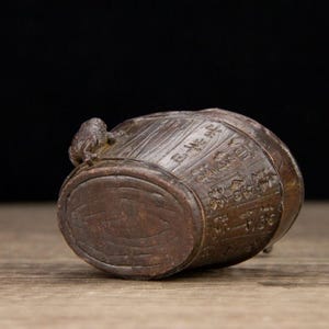 Puede incluir: Un peque&ntilde;o barril de madera antiguo con caracteres chinos tallados y una peque&ntilde;a figura de rana en la parte superior. El barril es marr&oacute;n con una superficie texturizada y un fondo oscuro.