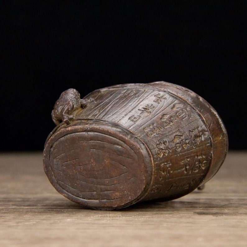 Puede incluir: Un peque&ntilde;o barril de madera antiguo con caracteres chinos tallados y una peque&ntilde;a figura de rana en la parte superior. El barril es marr&oacute;n con una superficie texturizada y un fondo oscuro.