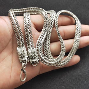 Puede incluir: Un collar plateado con un diseño de cadena trenzada. El collar presenta dos colgantes con cabeza de dragón y un cierre. La joya se muestra en una mano sobre un fondo oscuro.
