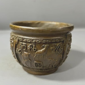 May include: A bronze-colored, round planter with embossed Chinese characters and images. The planter has a wide, open top and a decorative band around the rim. The sides feature detailed relief carvings of figures and symbols.