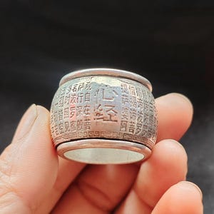 May include: A silver ring with engraved text. The ring has a cylindrical shape with a slightly flared top and bottom. The text appears to be in a foreign language, possibly Chinese or Japanese. The ring is held in a person's hand.