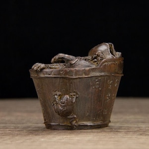 Puede incluir: Peque&ntilde;a escultura de madera marr&oacute;n de una rana relaj&aacute;ndose en un cubo de madera. La rana est&aacute; esculpida con detalle, con los brazos apoyados en el borde del cubo. Tambi&eacute;n hay una rana tallada en el lateral del cubo.