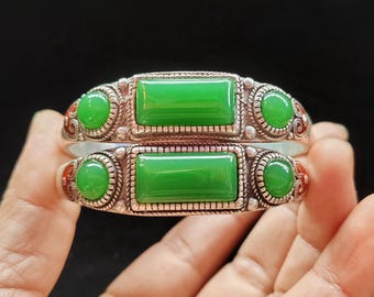 May include: Two silver bracelets with bright green rectangular and round jade stones. The bracelets have intricate silver detailing and red enamel accents. The bracelets are displayed against a dark background.