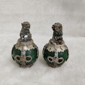 May include: Two decorative green and silver urns. Each urn features a detailed silver lion figure on top. The urns have a round shape with silver accents and dragon-like designs. The background is a soft, white fabric.