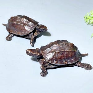 Op de afbeelding: Twee bronzen schildpadbeeldjes. De schildpadden hebben gedetailleerde schilden en staan ​​in een loophouding. De beeldjes staan ​​op een lichtgrijze ondergrond. De schildpadden zijn een decoratief item.